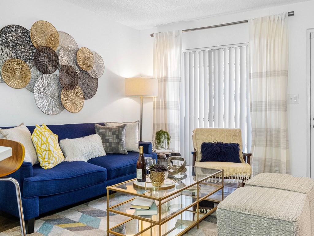 A living room with a blue couch, a glass coffee table, and a large decorative piece on the wall.